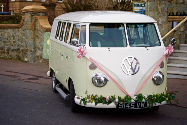 Ein weißer und pinker VW-Bus mit floralen Verzierungen, der vor einer Steinmauer mit Treppe und einem Gebäude im Hintergrund geparkt ist.