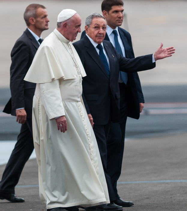 Pope Francis, wearing a white robe and cap, arrives at St. Peter's Square at the Vatican, accompanied by four men in black blazers and ties.