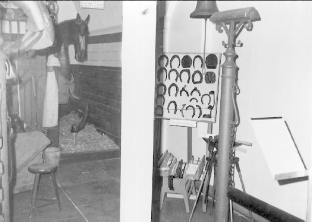 Schwarzes und weißes Foto eines Raums mit einem Pferd im Hintergrund, einer Person auf der linken Seite und verschiedenen Gegenständen, einschließlich eines Hockers und an der Wand angebrachten Rahmen, die auf einen Pferdestall hinweisen.