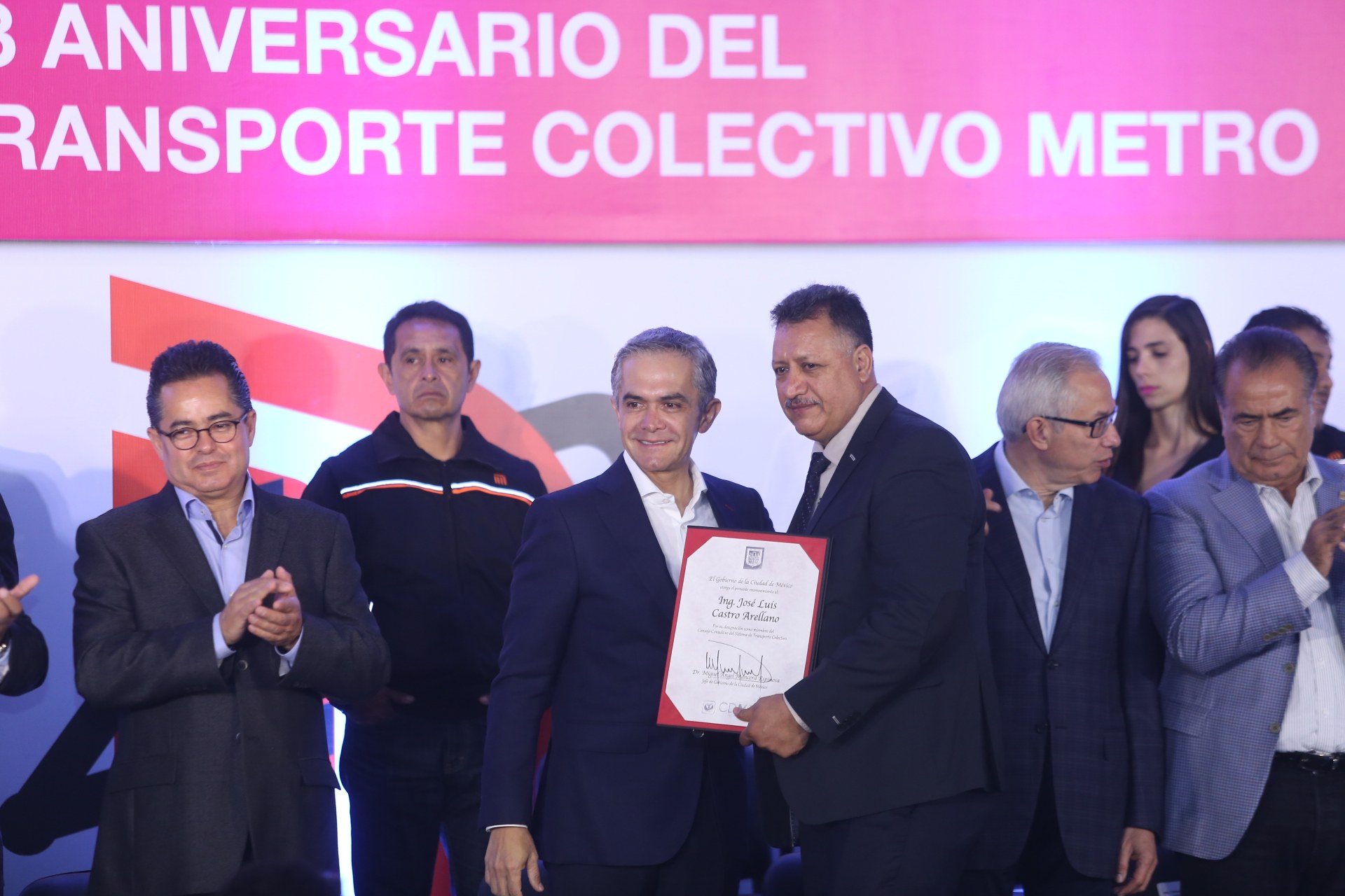 Three men on a stage holding a certificate, with a banner in the background reading "3 aniversario del transporte collectivo metro."