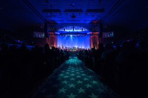 Ein großer Auditorium mit Sitzplätzen, einer Bühne mit Musikern und Überkopfbeleuchtung und Fernsehern, was auf einen Konzertsaal während der Nationalkonvention 2019 hinweist.