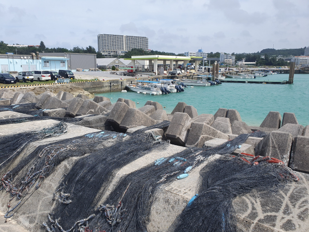 Ein großer Haufen Fischernetze auf einem Steg neben Wasser mit Booten, Fahrzeugen, Gebäuden, Bäumen, Pfählen und einem bewölkten Himmel im Hintergrund.