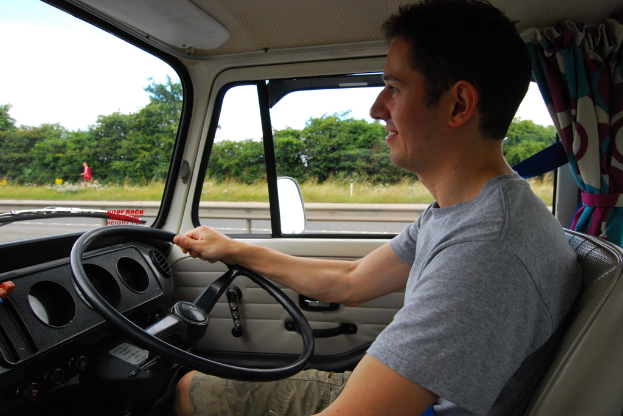 Eine Person in einem grauen T-Shirt und Shorts fährt ein Fahrzeug, hält das Lenkrad, mit Vorhängen sichtbar, während eine andere Person in einem roten Kleid hinten steht, mit Bäumen im Hintergrund.