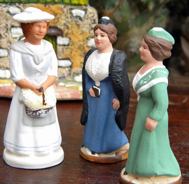 Drei Frauen-Skulpturen-Puppen sind auf einer Holzplatte mit Spielzeughäusern im Hintergrund angeordnet.