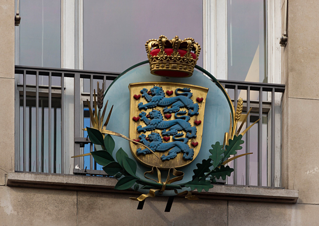 Wappen der britischen Königsfamilie auf einem Gebäude mit einer Krone und einem Blatt-Logo, Fenstern und Metallgittern im Hintergrund.