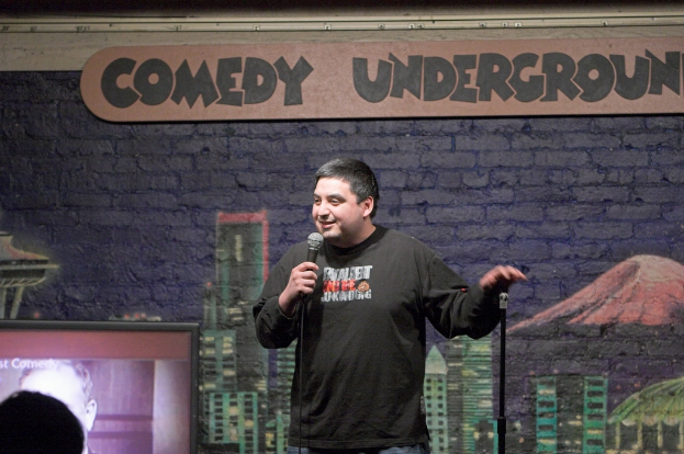 Ein Mann steht mit einem Mikrofon in der Hand und spricht, mit einem Bild an der Wand, einem Fernseher und Text an der Wand.