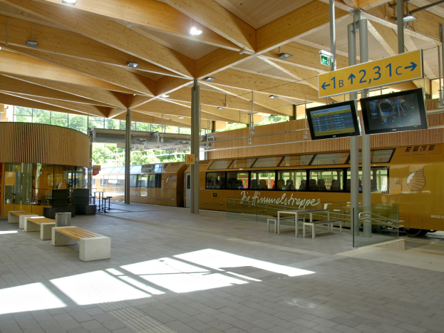 Ein belebter Bahnhof mit einem Zug auf den Schienen, umgeben von Bänken, Tischen, Pfosten, Schildern, Laternen und Bäumen und voller Menschen, die auf den Zug warten.
