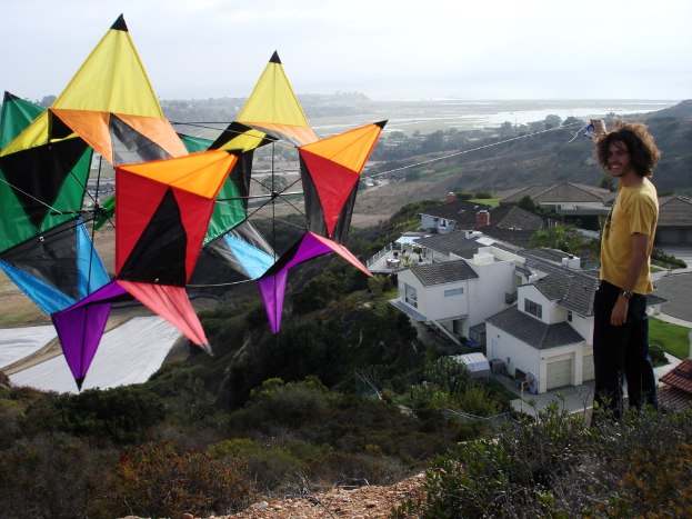 Ein Mann in einem gelben T-Shirt hält ein Seil, das an einem Kite befestigt ist, mit Häusern, Bäumen und dem Himmel im Hintergrund.