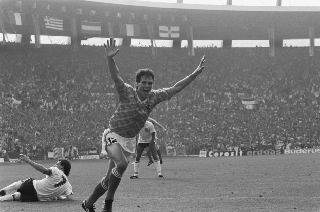 Schwarzes und weißes Foto eines Füßballspielers, der nach einem Tor in einem Stadion feiert, umgeben von Mannschaftskameraden und Mitarbeitern, mit Stadiontafeln, Fahnen und Masten im Hintergrund.
