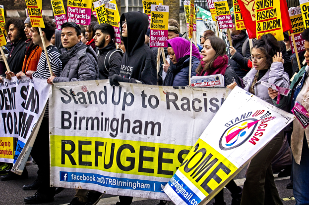 Group of people marching down a street holding protest signs and banners, with a building in the background.