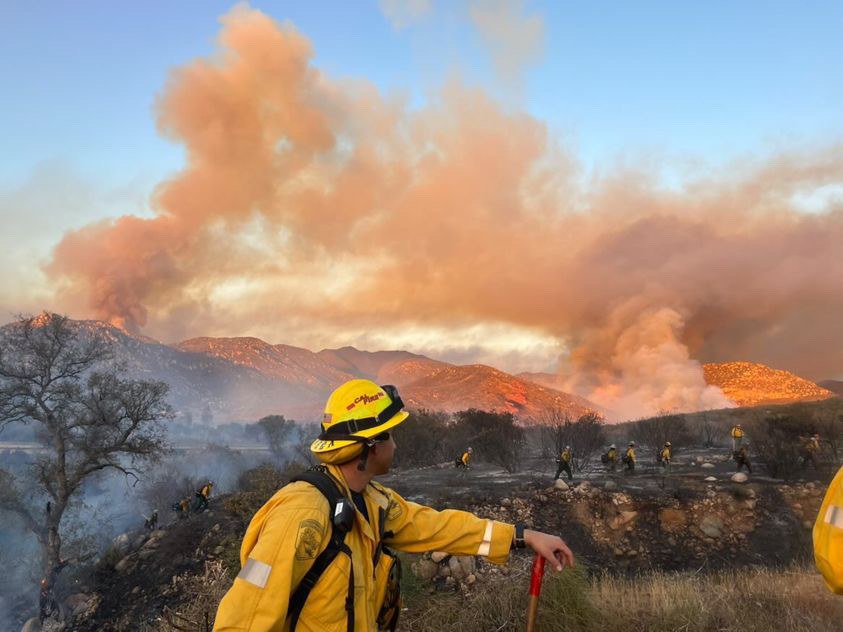 A firefighter in a yellow jacket and helmet stands in front of a large plume of smoke, holding a stick, with trees, hills, and a clear blue sky in the background.