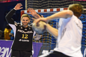 Zwei Männer spielen Handball vor einem Tor mit Netz und einem Banner mit Text im Hintergrund und einigen Menschen in einer Stadion.