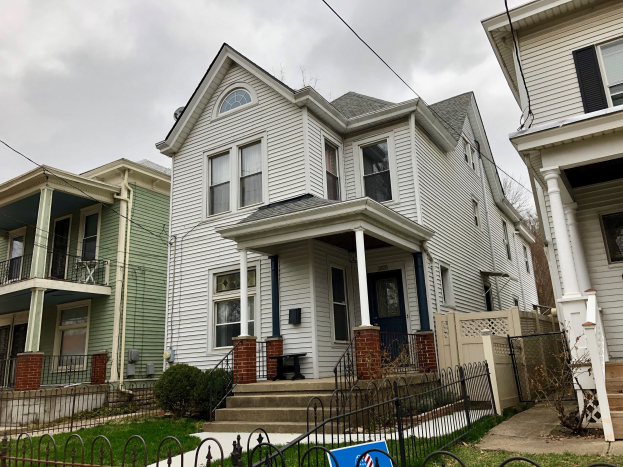 Reihe von Häusern mit Fenstern, Türen, Säulen, Geländern, Stufen und Dächern, umgeben von einem Metallzaun, Gras, Pflanzen, Bäumen und einem bewölkten Himmel, mit einem Mietschild unten, das die Adresse und Listen Details anzeigt.