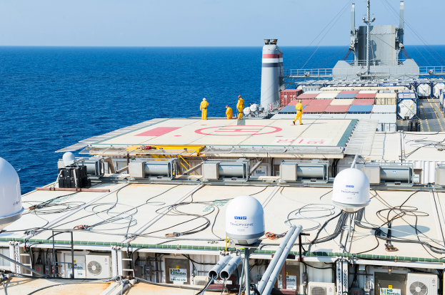 Eine Gruppe von Menschen auf einem großen Schiff im Meer, umgeben von Containern, Pfählen und Drähten, mit einem Offshore-Windpark im Hintergrund.