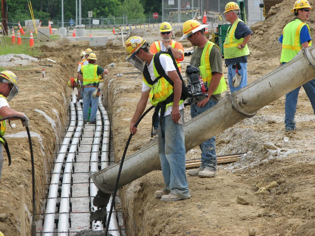 Eine Baustelle mit Arbeitern in Jacken und Helmen, Absperrgitter, Zaun, Bäume, Pfosten, Erde und Rohrleitungen im Hintergrund.
