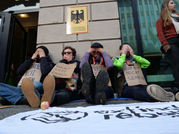 Eine Gruppe von Menschen sitzt vor einem Gebäude auf dem Boden, hält Schilder hoch, mit einer Fahne und einem Apfel auf der Straße, und eine Frau steht rechts daneben und sieht besorgt aus.
