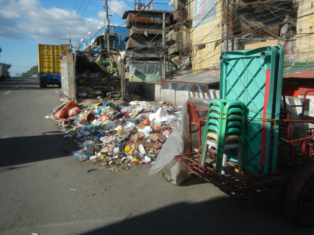 Ein Lastwagen steht neben einem Müllhaufen auf einer Straße, mit einem Wagen mit Plastikstühlen rechts daneben und Gebäuden, Strommasten, Bäumen und einem bewölkten Himmel im Hintergrund.