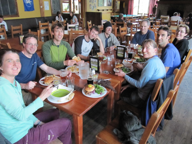 Eine Gruppe von Menschen sitzt um einen Tisch mit Tellern, Schüsseln, Gläsern, Flaschen, einem Wasserglas, Karten, Tüchern und Essen, lachend, mit einer Jacke auf einem der Stühle, während einige weitere Personen an anderen Tischen sitzen, mit einem Banner, Bildern und Postern an der Wand und einem Fenster mit Vorhängen.
