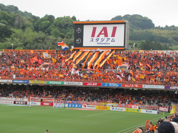 Ein Fußballspiel wird in einem Stadion mit einer großen Menge, grünem Rasen, einem Torpfosten, Bannern, Fahnen, einem großen Bildschirm, Bäumen und einem klaren blauen Himmel gespielt.