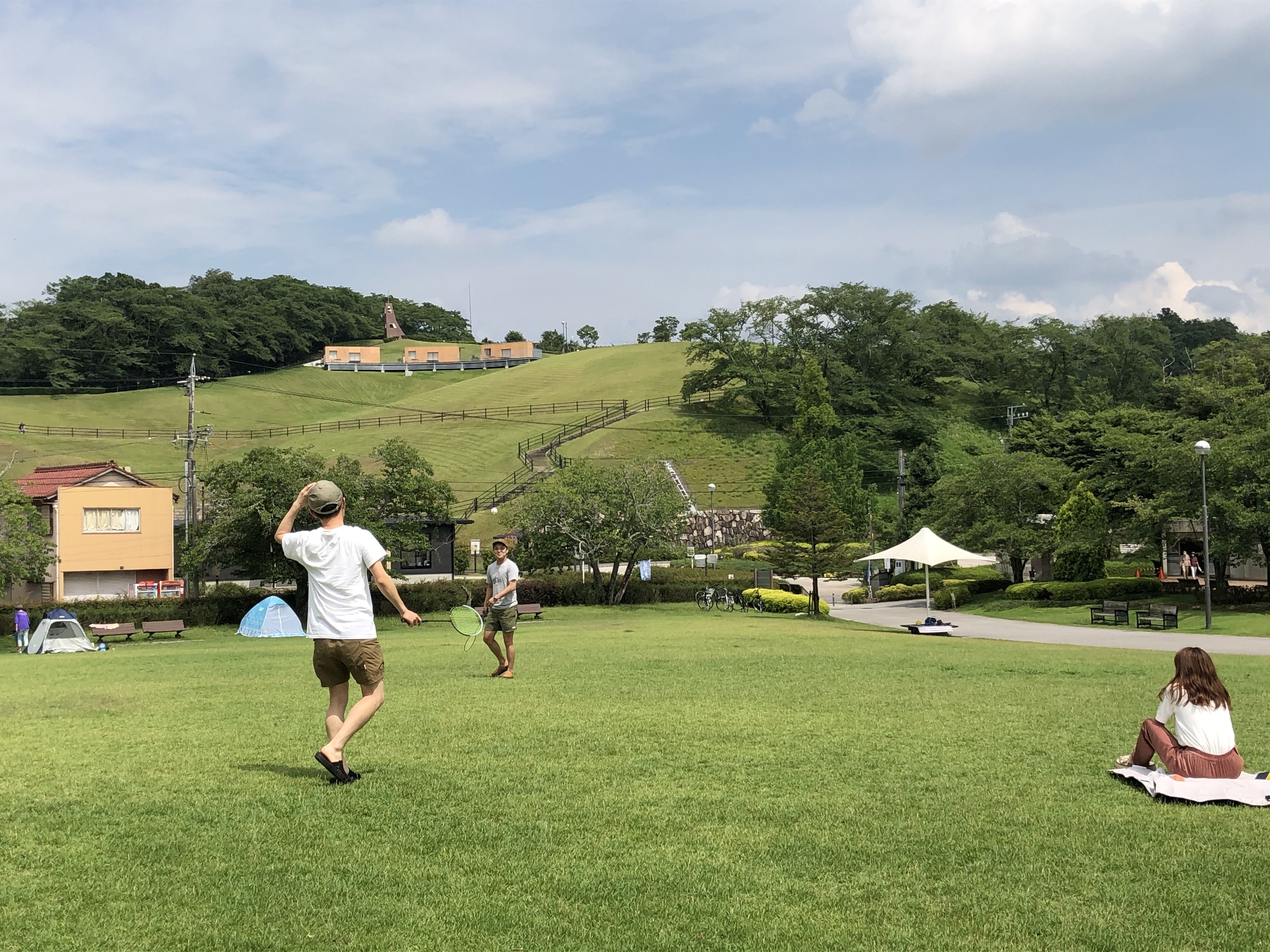 Gruppe von Menschen, die Badminton in einem Park spielt, mit einem Mann, der einen Schläger hält und auf Gras in der Nähe von Zelten und Gebäuden sitzt, mit Hügeln und einem bewölkten Himmel im Hintergrund.