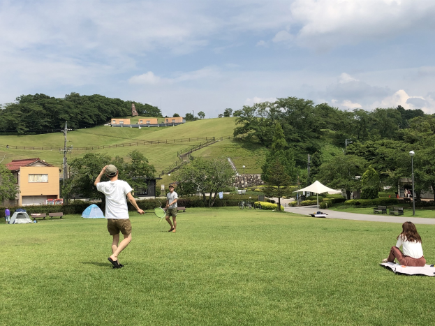 Gruppe von Menschen, die Badminton in einem Park spielt, mit einem Mann, der einen Schläger hält und auf Gras in der Nähe von Zelten und Gebäuden sitzt, mit Hügeln und einem bewölkten Himmel im Hintergrund.