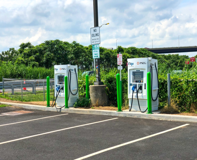 Ladestation für Elektrofahrzeuge auf einem Parkplatz mit umgebender Infrastruktur und einer Brücke im Hintergrund.