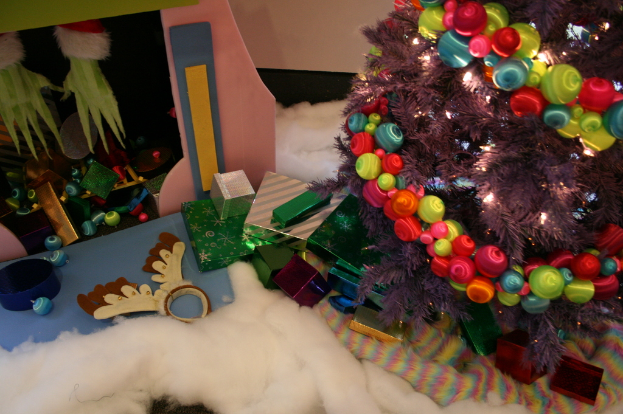 Eine Innenaufnahme mit einem geschmückten Weihnachtsbaum auf der rechten Seite, Geschenkboxen und Dekorationen in der Mitte und einer Hütte mit Dekorationen oben links in der Ecke, mit Watte und Wolle unten.