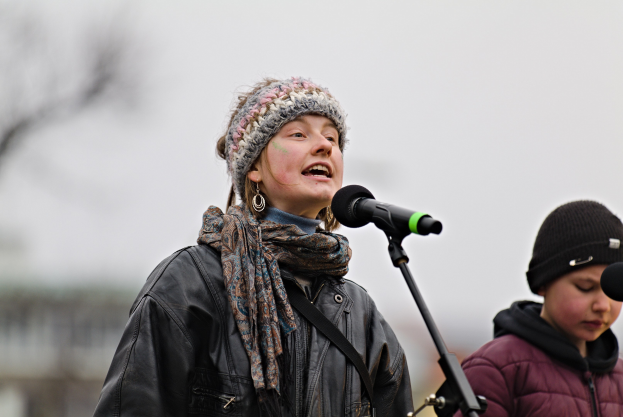 Eine Frau in einer schwarzen Jacke, einem Schal und einer Mütze singt in ein Mikrofon auf der Bühne, neben einer Person in einer marinefarbenen Jacke und einer schwarzen Mütze, während eine unscharfe Menge zusieht.
