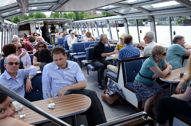 Boot-Innenraum mit Sitzreihen, vor denen Gläser und Schüsseln auf Tischen stehen, einer Tasche auf dem Boden, Wasser im Hintergrund, Gebäuden auf der rechten Seite und kleinen Pflanzen über dem Boot.