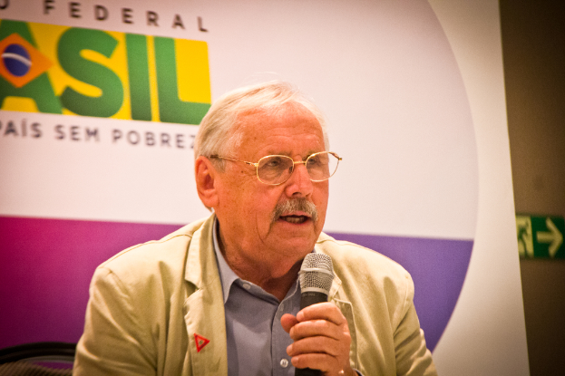 Eine Person, die in ein Mikrofon spricht und im Hintergrund ein Banner mit der Aufschrift 'Bundes' zu sehen ist.