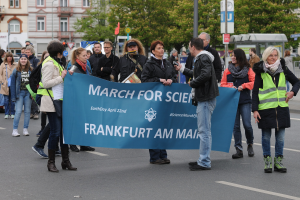 Gruppe von Menschen marschiert die Straße entlang und hält ein "March for Science Frankfurt am Main"-Schild mit Bäumen, Pfosten, Schildern und Gebäuden im Hintergrund unter einem klaren blauen Himmel.