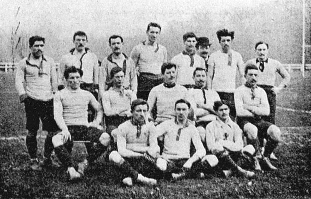 Ein Schwarz-Weiß-Foto einer Fussballmannschaft aus den 1920er Jahren, mit einigen stehenden und einigen sitzenden Spielern auf dem Boden, mit Pfählen, Bäumen und einem klaren Himmel im Hintergrund.