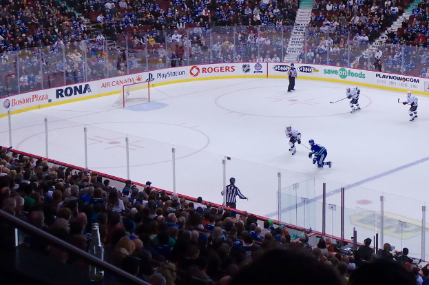 Vier Männer beim Eishockey auf einem Eisstadion mit zwei in der Nähe des Tors und Zauns stehenden Männern, Zuschauern auf Stühlen im Hintergrund und einer Treppe sichtbar.