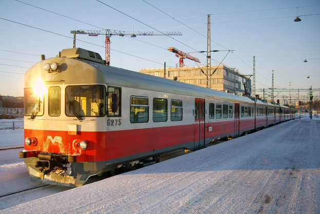 Ein roter und weißer Zug fährt auf schneebedeckten Schienen, mit Strommasten und Kabeln daneben, Gebäuden und Bäumen im Hintergrund unter einem klaren Himmel.