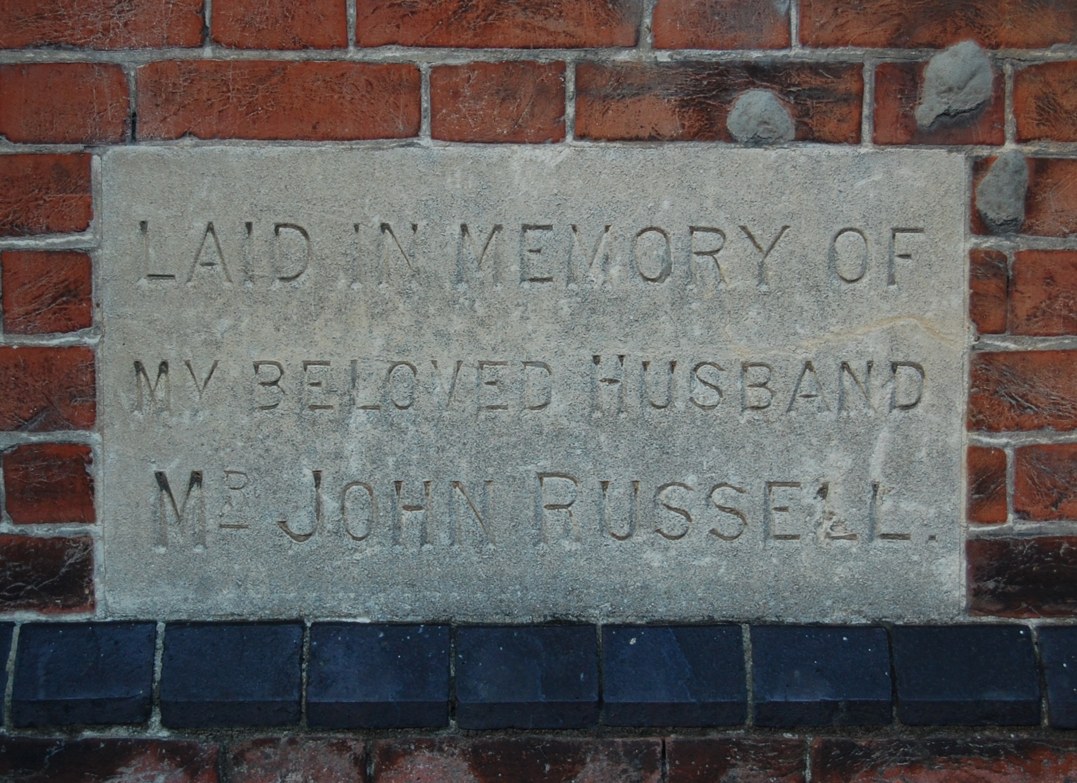 Stone plaque mounted on a brick wall reading "Laid in Memory of My Beloved Husband".