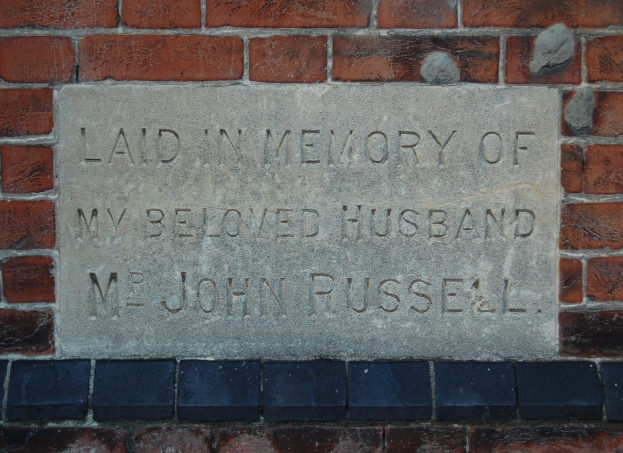 Stone plaque mounted on a brick wall reading "Laid in Memory of My Beloved Husband".