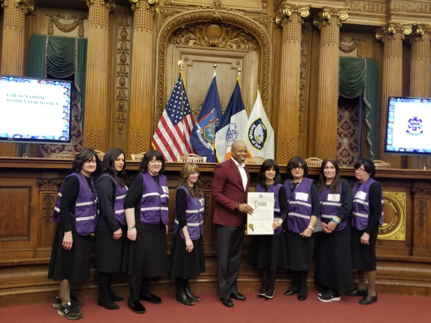 Gruppe von Menschen vor Flaggen mit einem Mann in der Mitte, der ein Zertifikat hält, flankiert von zwei Bildschirmen, vor einer hölzernen Wand mit kunstvollen Schnitzereien und Vorhängen, wahrscheinlich eine Stadtratssitzung.