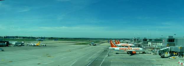 Eine Luftaufnahme eines Flughafens mit Flugzeugen, Gebäuden, Masten, Fahrzeugen, Bäumen, einer Straße unten und einem Zaun im Vordergrund.