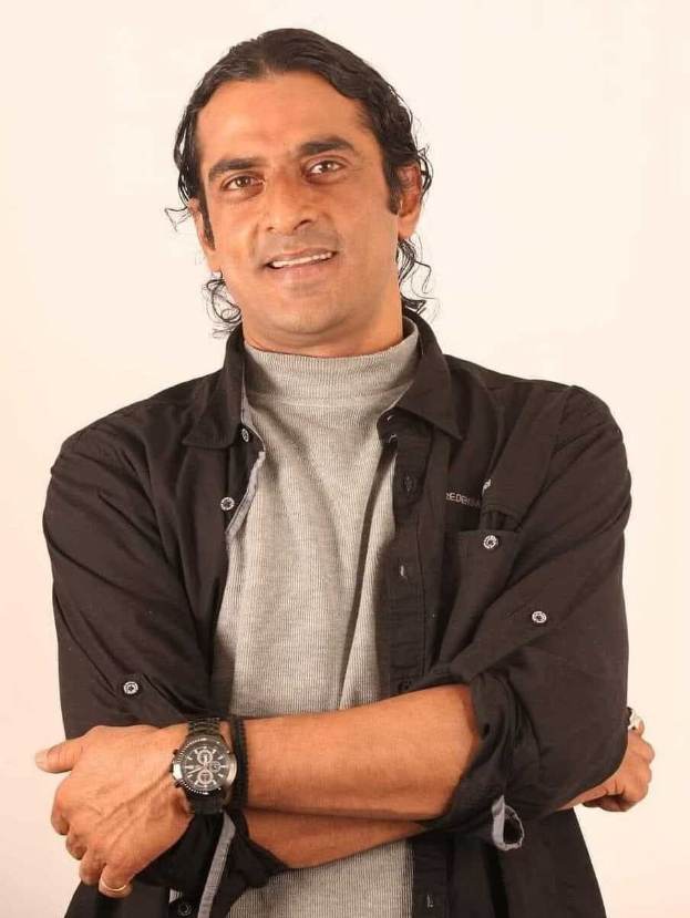 Ein Mann in einem schwarzen und aschfarbenen Kleid mit überschlagenen Armen, der eine Uhr tr├Ągt und gegen einen weißen Hintergrund steht, der nachdenklich wirkt.