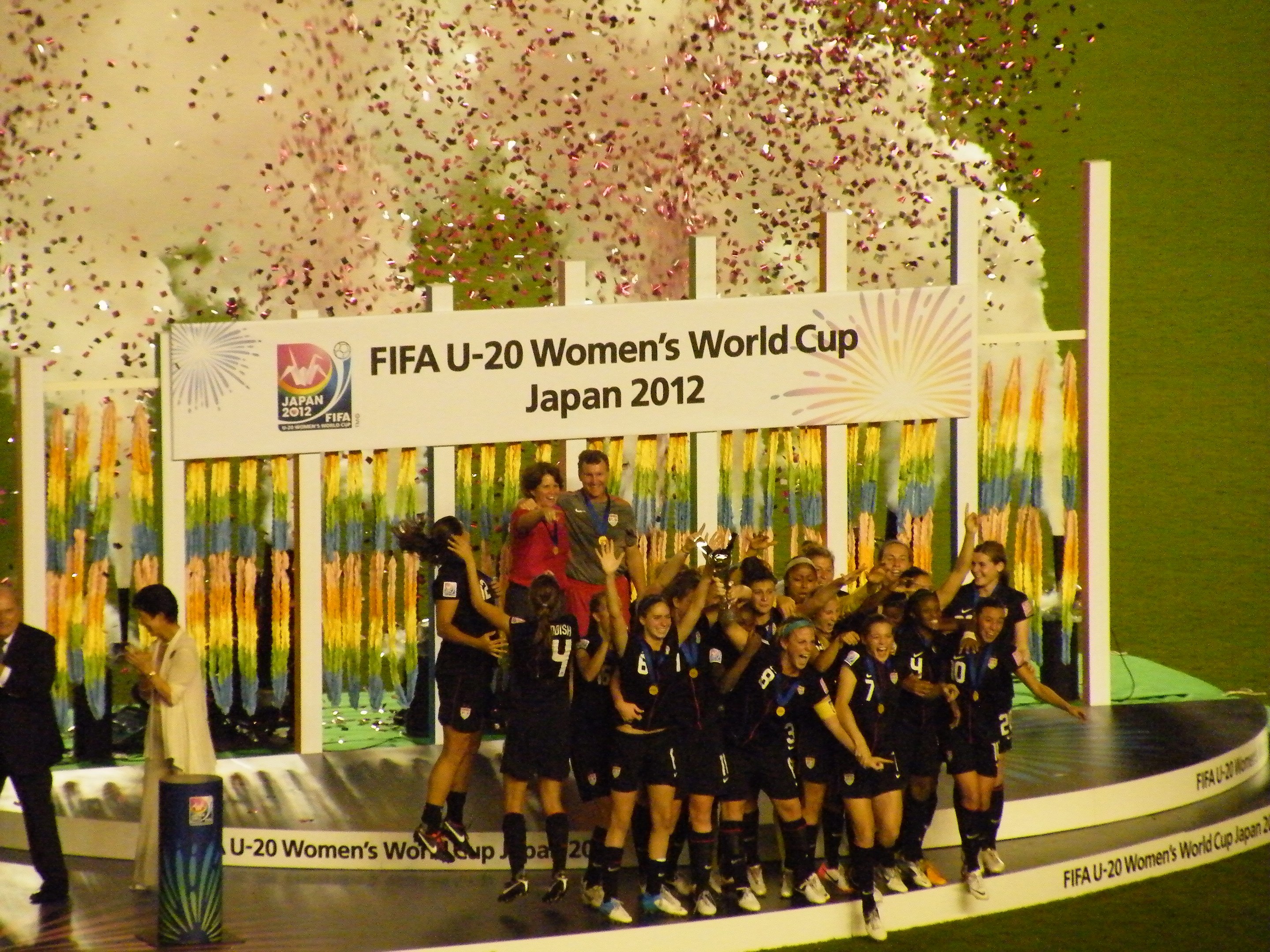 Gruppe von Frauenfußballspielerinnen in schwarzen Uniformen, die auf einer Bühne umgeben von einer Menge feiern, mit einem Hintergrundboard und dekorativen Gegenständen auf Gras.