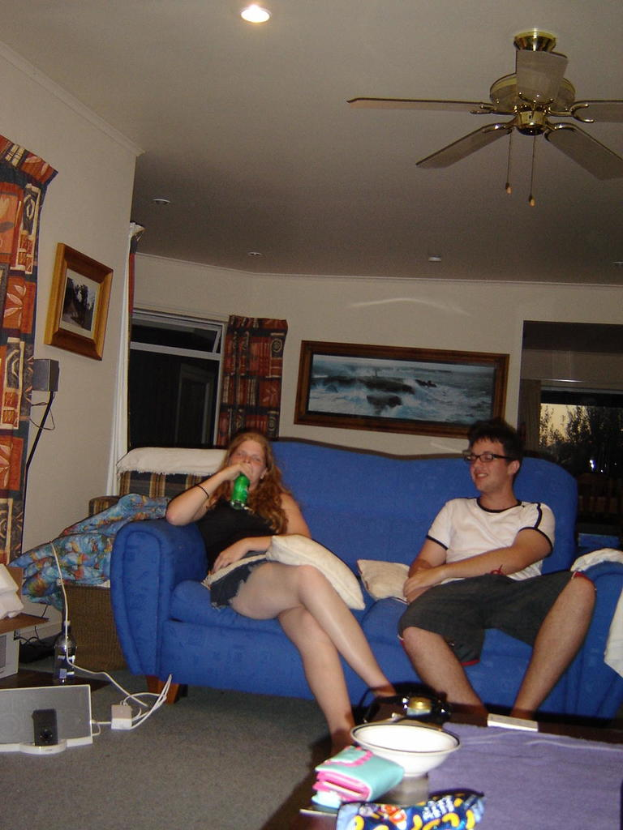 Eine Frau sitzt auf einem Sofa und hält eine Flasche in der Hand, neben ihr ein Mann; auf dem Tisch liegen ein Kissen, eine Schößel, eine Geldbörse und ein Tuch, an der Wand hängt ein Rahmen, Überkopfleuchte, Ventilator und Vorhang im Hintergrund, mit einem Baum im Freien.