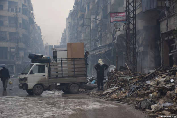 Ein Lkw fährt auf einer Straße neben einem Schutthaufen, mit Menschen auf der Straße, einer hält einen Schirm, und Gebäuden, Pfählen und Schildern mit Text im Hintergrund bei klarem Himmel.