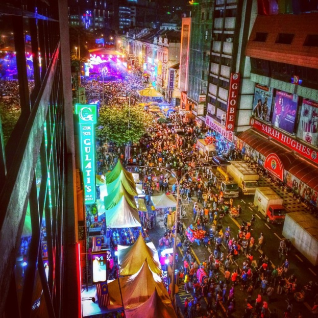 Eine belebte Karnevalsszene mit einer großen Menge auf der Straße, umgeben von Zelten, Geschäften, Gebäuden und Bannern, mit hell erleuchteter Fläche im Hintergrund.