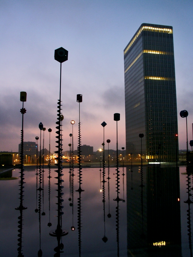 Mehrere Pole und Gebäude mit Lichtern unter einem bewölkten Himmel, mit Spiegelungen der Pole sichtbar.