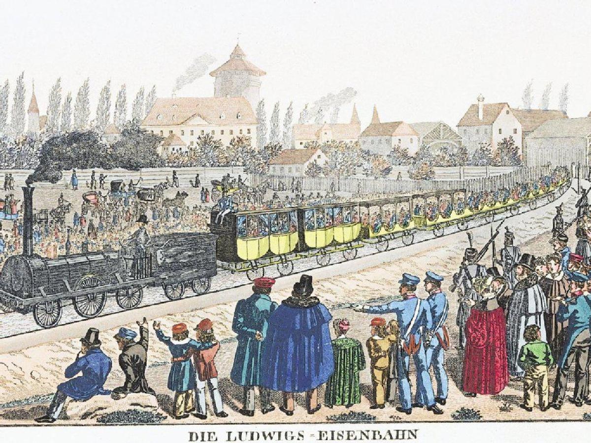 Eine Gruppe von Menschen in der Nähe eines Zuges auf einem Gleis, einige im Zug und andere auf dem Boden, vor Gebäuden und Bäumen im Hintergrund, mit dem Text "Ludwigs-Eisenbahn, der erste Bahnhof in Deutschland" unten.