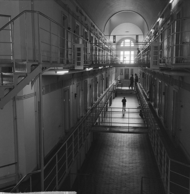 Schwarz-weißes Foto eines Mannes, der einen langen Gefängnishof mit Gittern, Türen und Lampen sowie ein Fenster im Hintergrund entlanggeht.