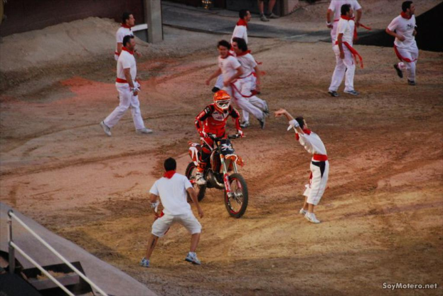Eine Person in rotem Kleid und Helm fährt ein Motorrad in der Mitte eines Platzes, umgeben von Jungen in weißen Kleidern, die laufen.