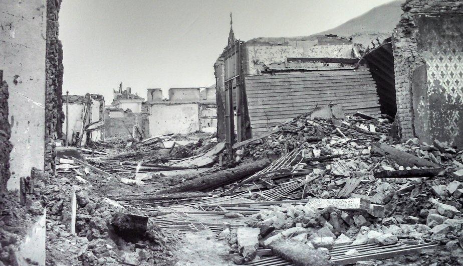 Ein Schwarz-Weiß-Foto eines schwer beschädigten Gebäudes auf einer Straße, mit Trümmern wie eingestürzten Wänden, Holzstöcken und Steinen, vor einem Hügel und dem Himmel.