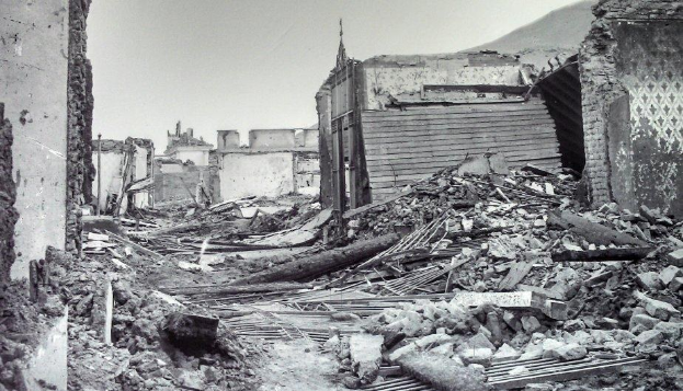 Ein Schwarz-Weiß-Foto eines schwer beschädigten Gebäudes auf einer Straße, mit Trümmern wie eingestürzten Wänden, Holzstöcken und Steinen, vor einem Hügel und dem Himmel.