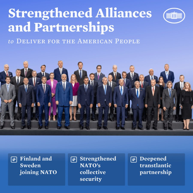 Gruppe von Menschen in Anzügen auf einer Bühne mit dem Text "Stärkere Allianzen und Partnerschaften, um für das amerikanische Volk zu liefern" oben und unten.